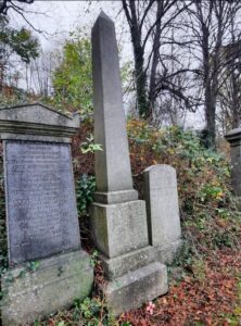 Monument to Gunner John Buchanan Monteith. Obelisk shaped. Cross with red Scottish poppy placed in front of monument amongst autumn leaves.  